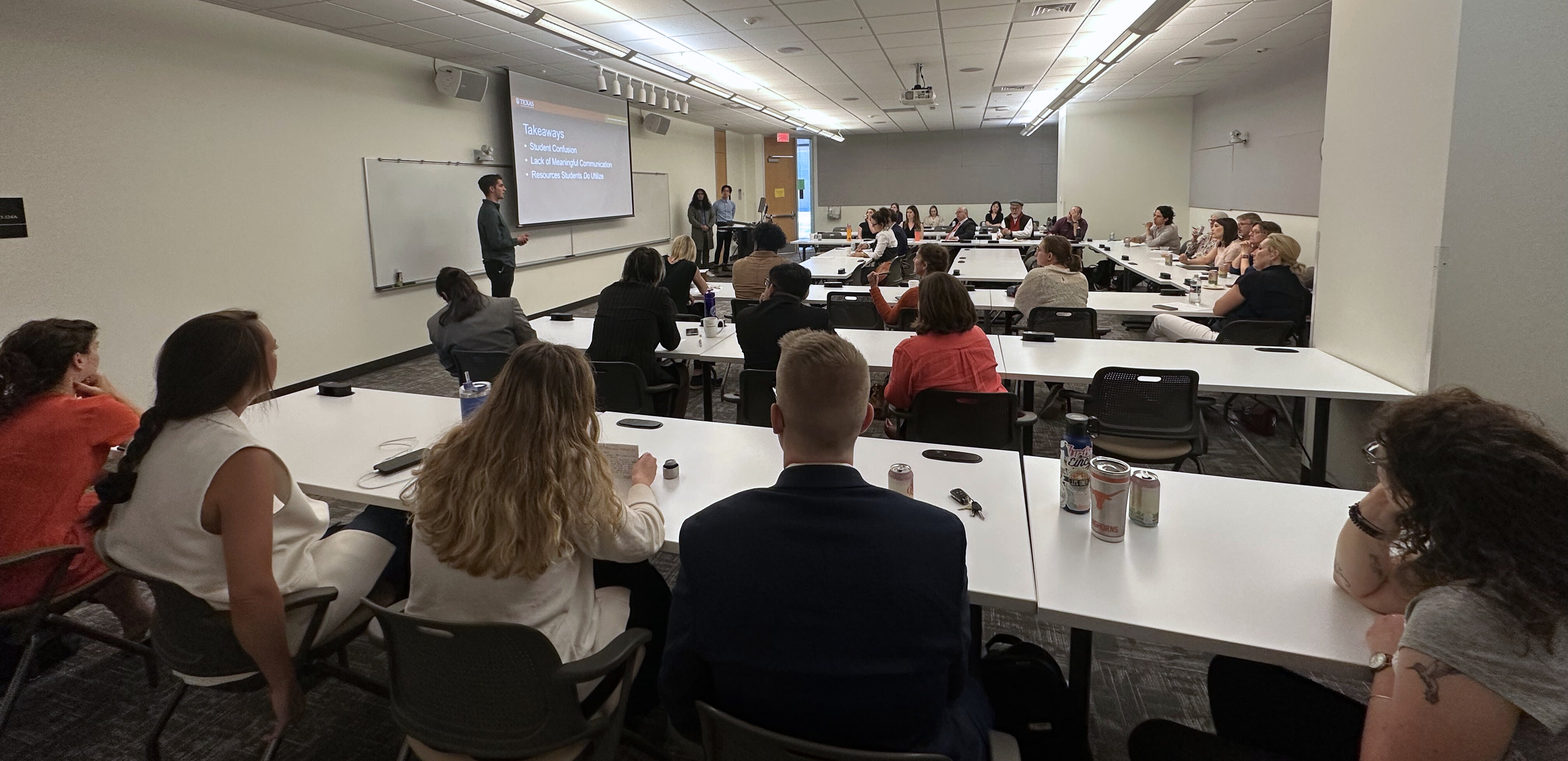 A group of students and guests stit in a classroom looking at students presenting at the front of the classroom