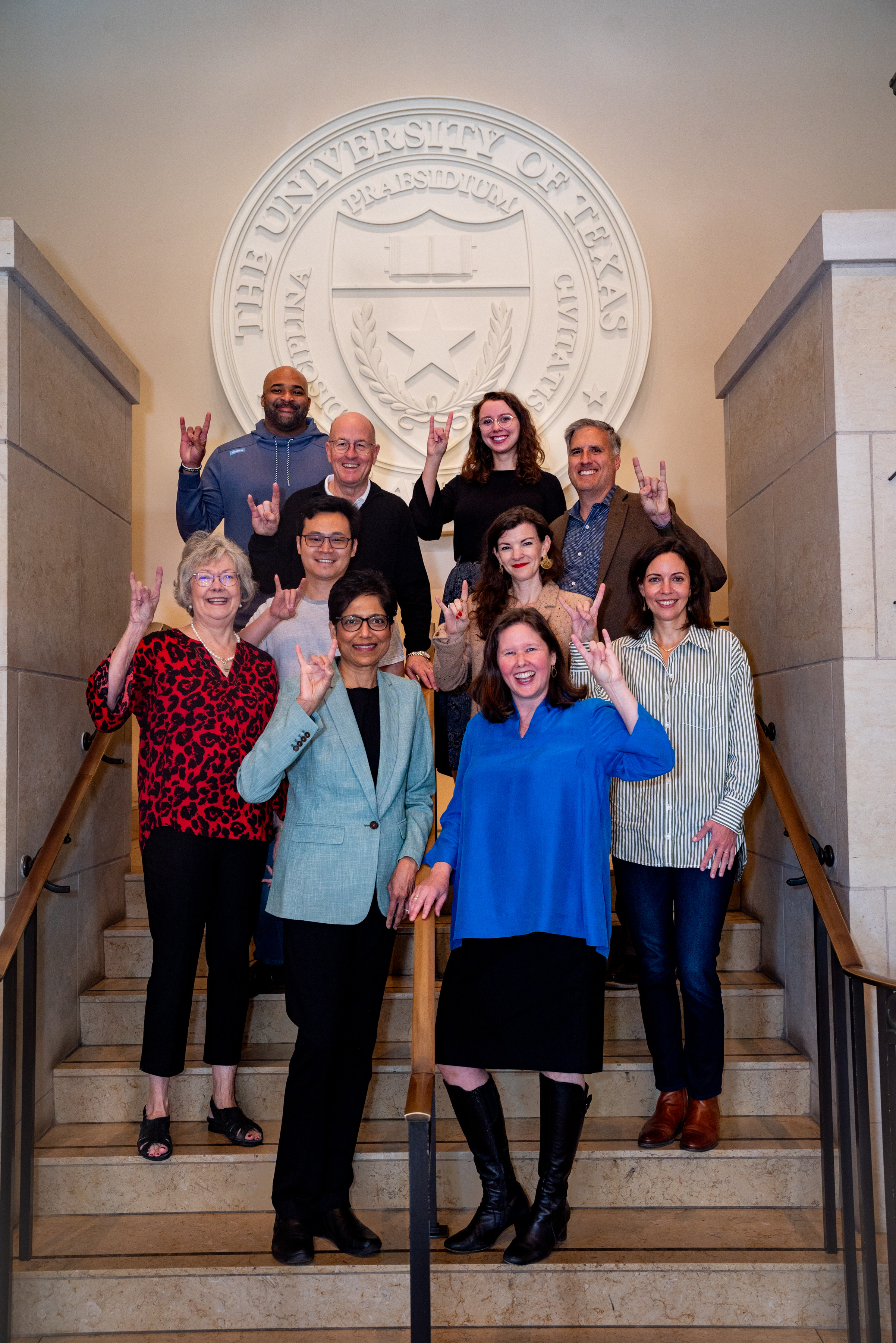 Group photo of RGK faculty in staff on the stairs. The group is standing in front of a large UT Austin seal and doing the "Hook 'Em" with their handsand 