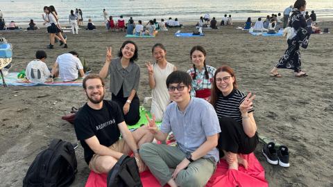 Zander Mintz sits with five other people on a blanket on the beach at sunset. They are smiling at the camera and making peace signs with their fingers.