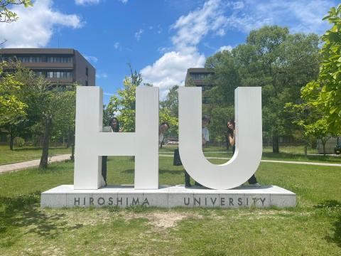 Neema stands with two other people behint giant letters "HU" on the Hiroshima University campus.