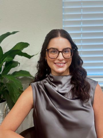 Headshot of Helaina, indoors in front of a potted plant. Helaina has long dark hair and glasses.