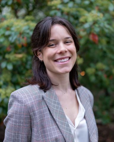 Headshot of Sydney, wearing a blazer and standing outside in front of greenery. Sydney has short brown hair.