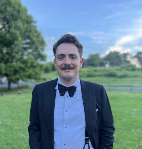 Headshot of Dylan standing outside in a green field. Dylan has short brown hair and a mustache, and is wearing a suit with a bowtie.