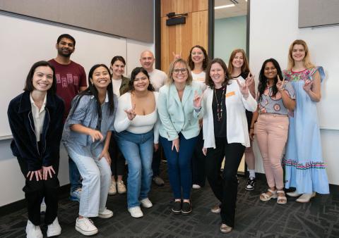 A group of students, staff, and alumni standing in a classroom smiling and holding up the Hook 'Em hand sign. 