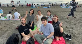 Zander Mintz sits with five other people on a blanket on the beach at sunset. They are smiling at the camera and making peace signs with their fingers.