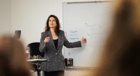 A Photo of Suzanne Smith standing in front of a whiteboard, tlaking to the classroom of students.