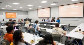 Photo of a classroom with students sittin gin rows. Three panelists and a student moderator are sitting in a row at the front of the classroom.
