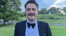 Headshot of Dylan standing outside in a green field. Dylan has short brown hair and a mustache, and is wearing a suit with a bowtie.