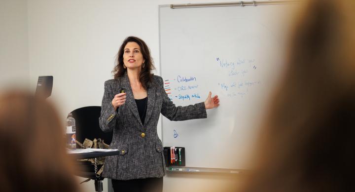 A Photo of Suzanne Smith standing in front of a whiteboard, tlaking to the classroom of students.